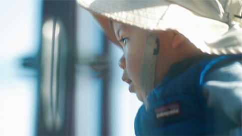 A baby wearing a hat and jacket stares out a window.